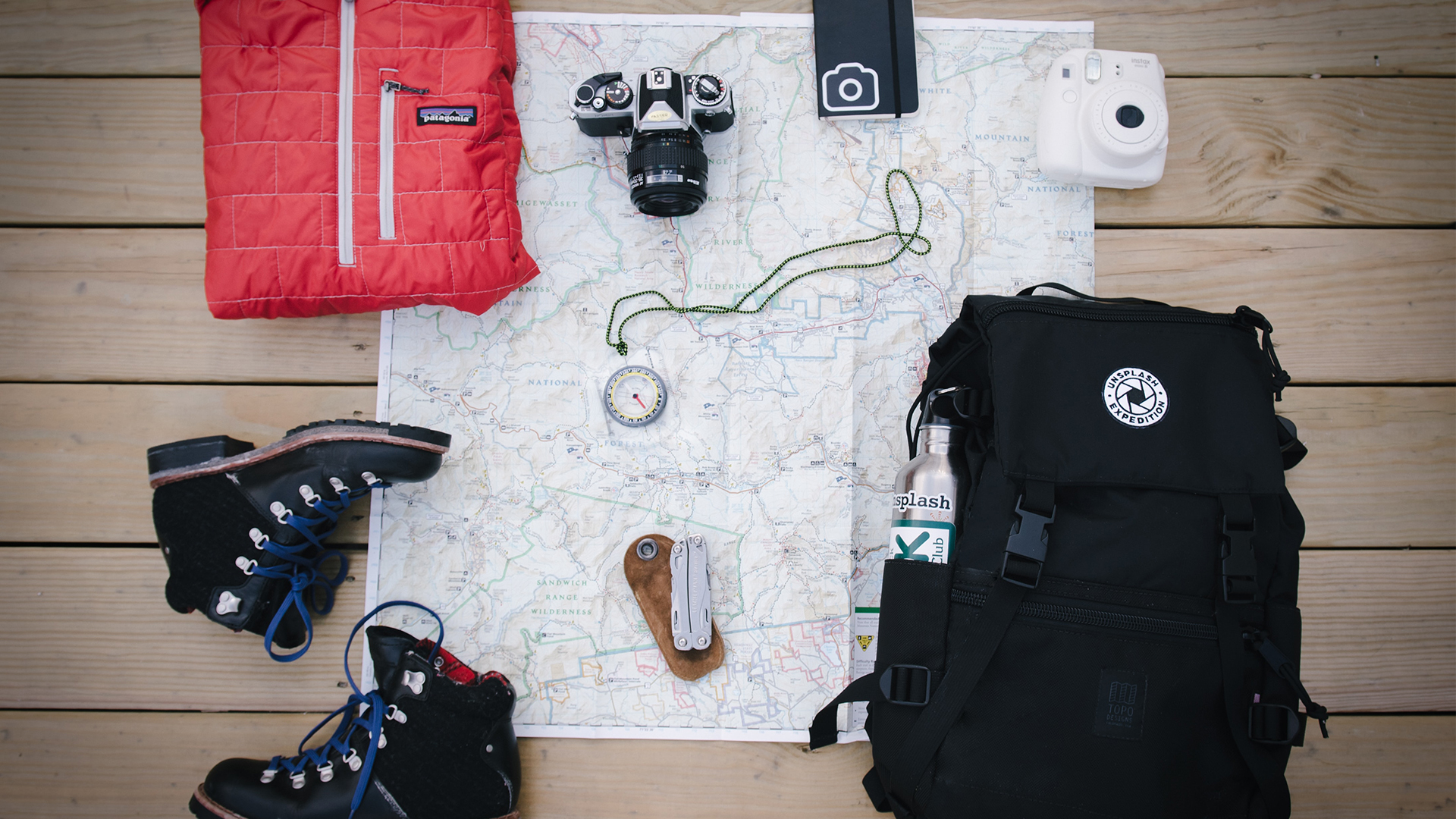 A red jacket, boots, two cameras, a notebook, compass, swiss army knife and backpack scattered on top of a map on the floor.