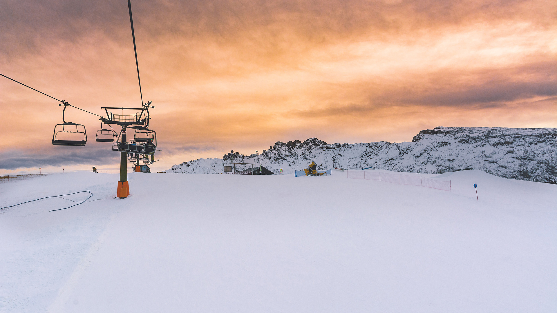 Snow covered ski field with ski lift on left hand side, and snow covered mountain in the background against a sunset.